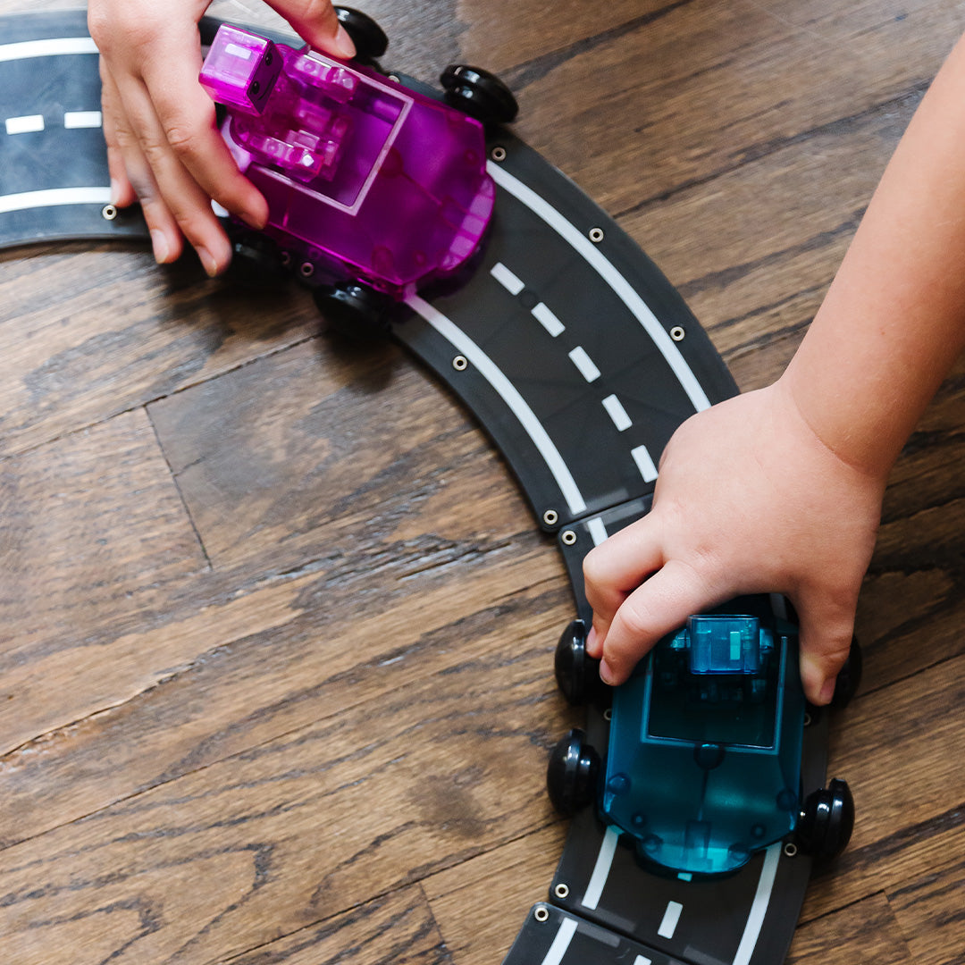 Twee kinderhanden duwen een paarse en een blauwe speelgoedauto over een gebogen weg van Magna-Tiles magnetische tegels.
