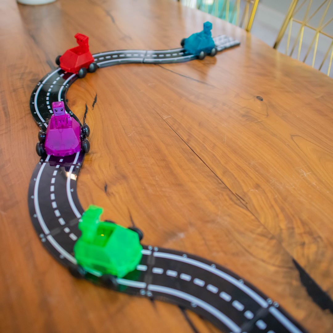 Een kronkelende weg van Magna-Tiles magnetische wegtegels met vier kleurrijke speelgoedauto's op een houten tafel.