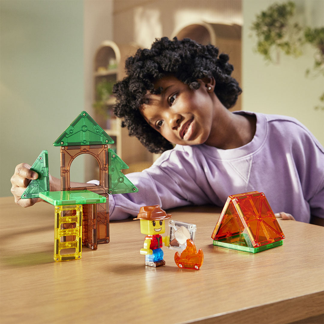 Een lachend kind speelt met de Magna-Tiles Camp Out set, bestaande uit een boomhut, een tent, een figuurtje en een kampvuur.