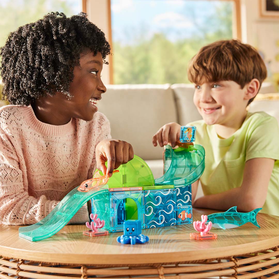 Twee kinderen spelen lachend met de Magna-Tiles Coral Reef magnetische bouwset op een houten tafel.