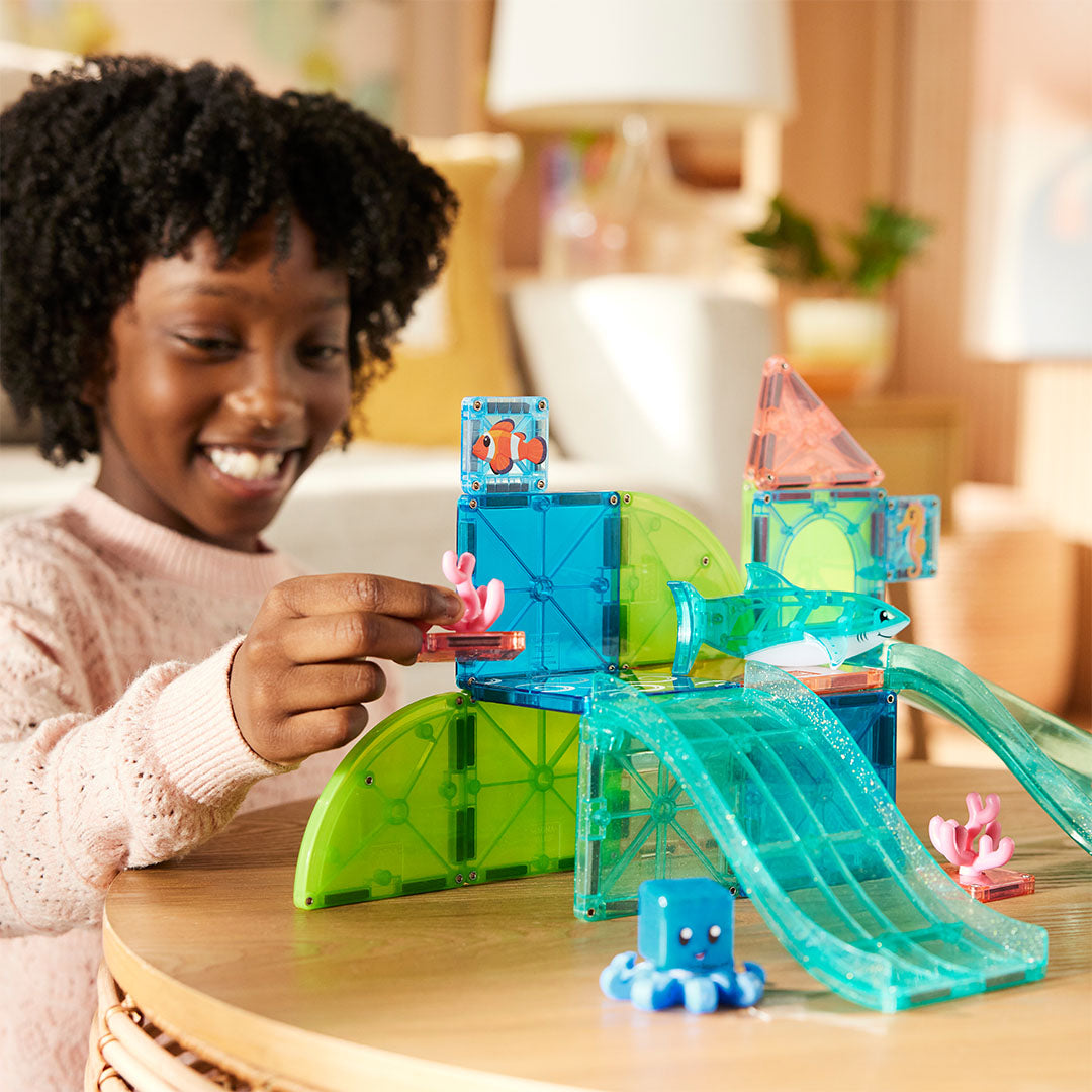 Een lachend meisje speelt met de Magna-Tiles Coral Reef magnetische bouwset en houdt een koraalstuk vast.