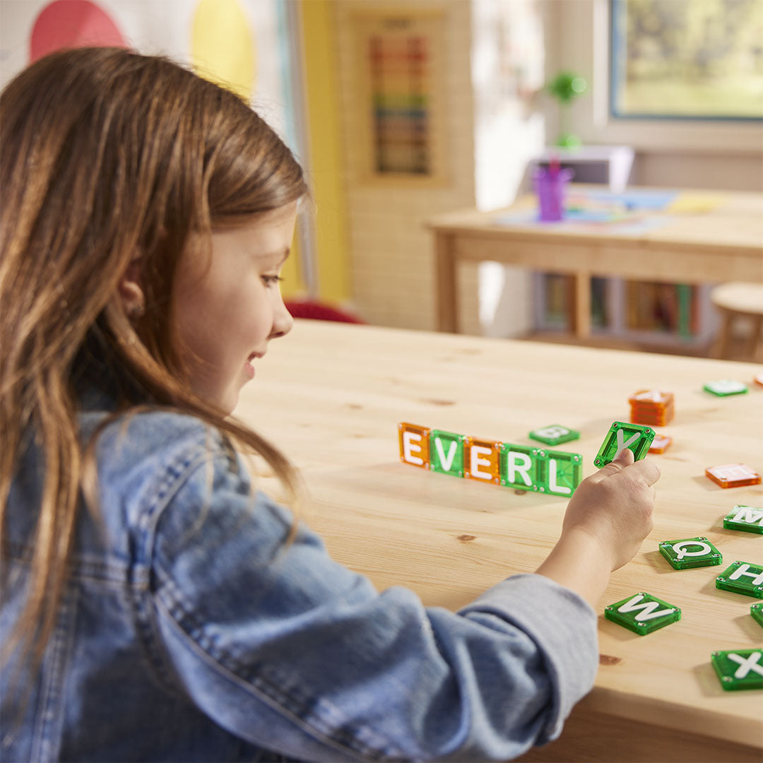 Een meisje legt Magna-Tiles magnetische lettertegels op een houten tafel om woorden te vormen.
