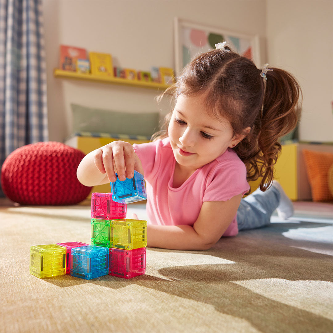 Een jong meisje bouwt een toren met kleurrijke Magna-Tiles magnetische bouwblokjes op de vloer.