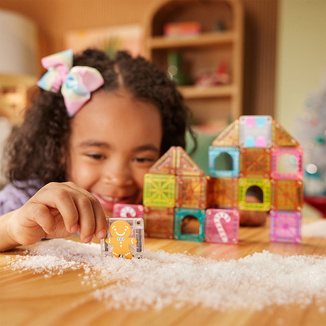 Een kind speelt met Magna-Tiles magnetische tegels, waarbij ze een koekjesman-tegel vasthoudt voor een gebouwd peperkoekhuis in een besneeuwde omgeving.