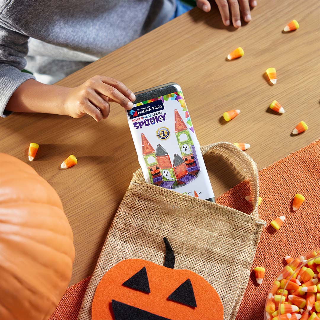 Handen plaatsen de Magna-Tiles MicroMags Travel Spooky bouwset verpakking in een Halloween-tas, omringd door snoep en een pompoen.