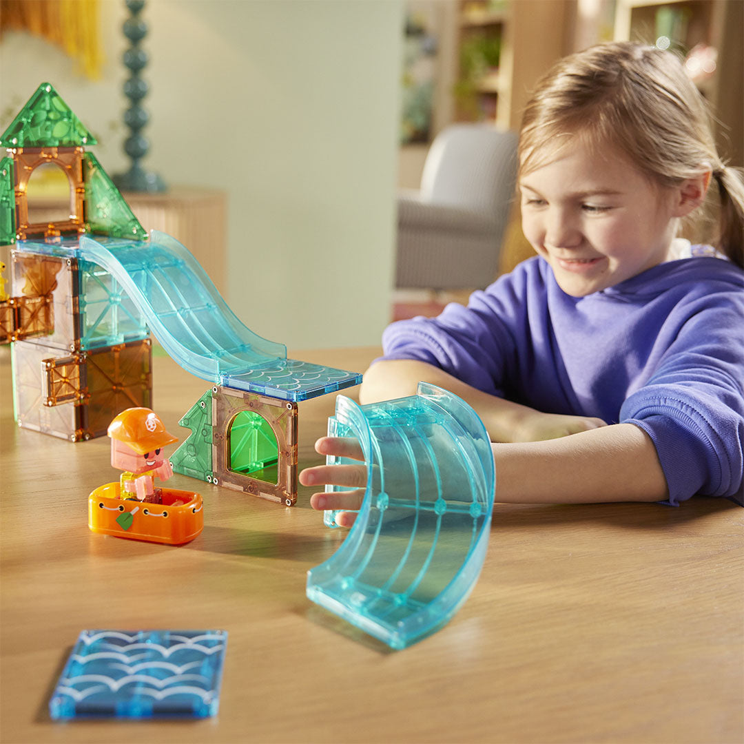 Een lachend meisje speelt met de Magna-Tiles Raging Rapids bouwset, een boomhut met glijbanen en een figuur in een bootje.