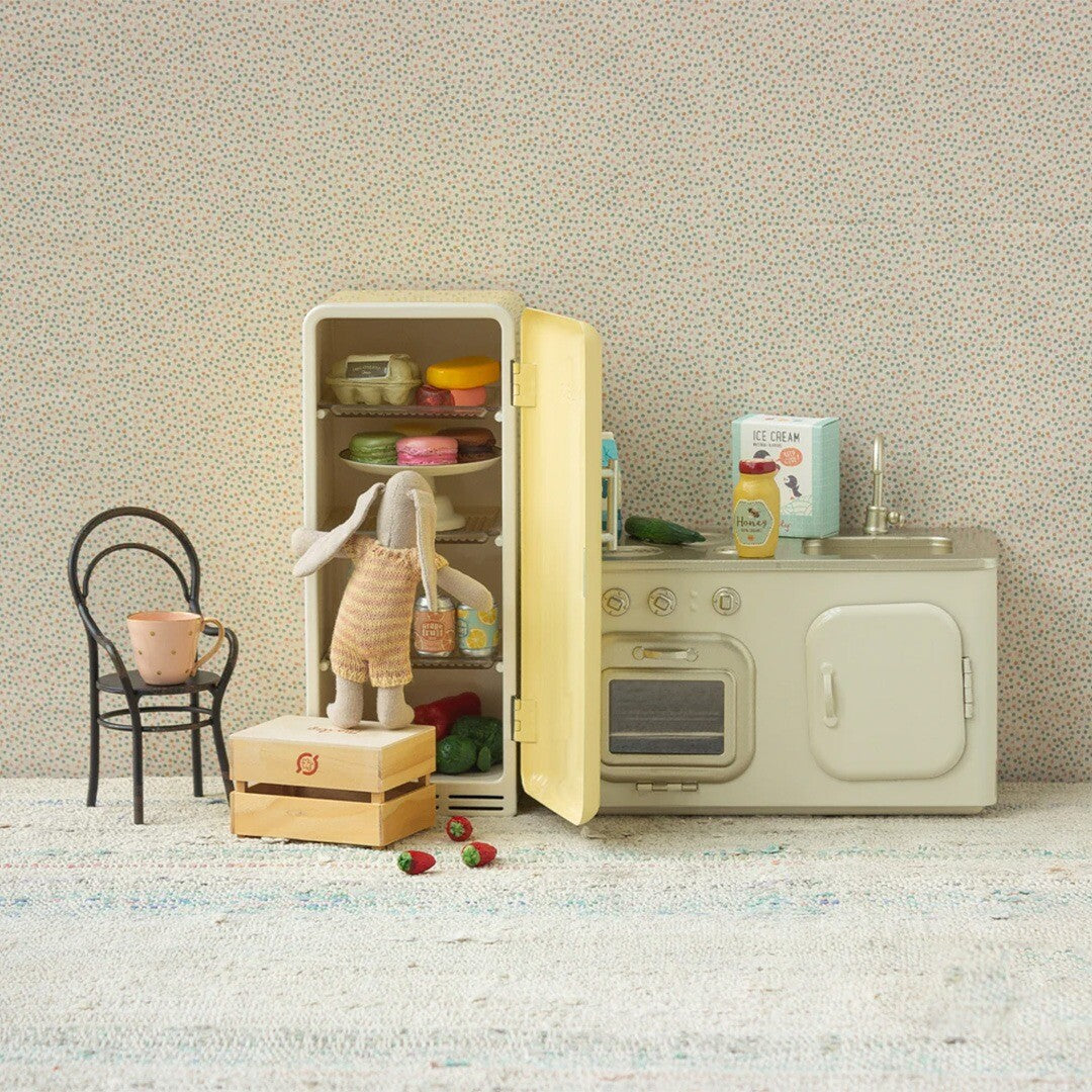 Maileg poppenhuis koelkast met open deur, gevuld met speelgoedvoedsel, naast een keukenblok en een konijnpop.