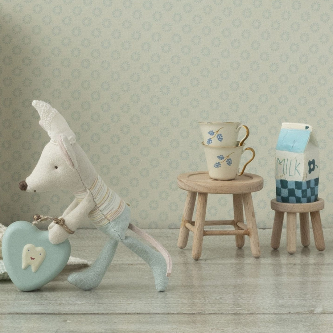 Een Maileg speelgoedmuisje trekt een hartvormig tandkussentje, met twee houten poppenhuis krukjes en miniatuur servies op de achtergrond.