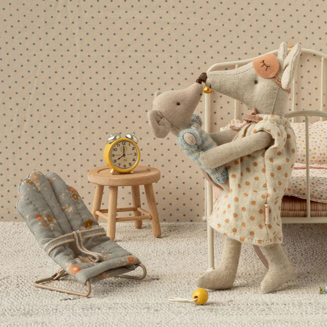 Een Maileg moedermuispop in een beige nachtjapon met stippen, die een babymuispop vasthoudt naast een bed en een poppenstoeltje.
