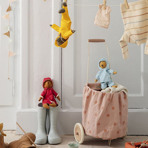 Maileg collectie met een teddybeer pop in rode regenjas en hoed, zittend op laarzen, naast een winkelwagentje met een andere pop.