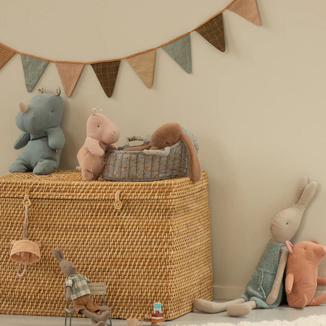 Maileg poppenreiswieg met een bruine konijn knuffel erin, staand op een rieten kist tussen andere knuffels.
