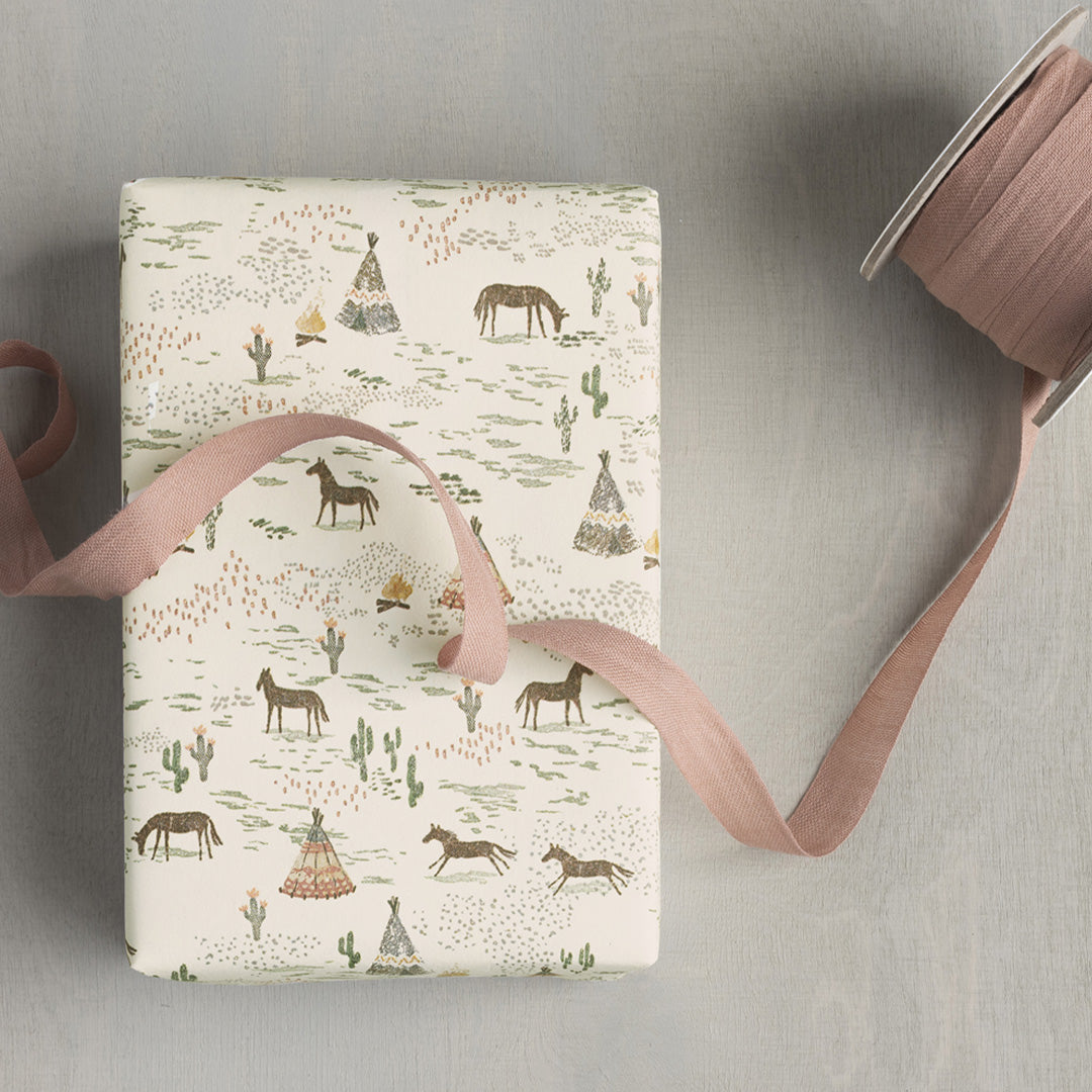 Maileg inpakpapier met paarden en tipi's, deels om een cadeau gewikkeld, met een rol roze lint ernaast.