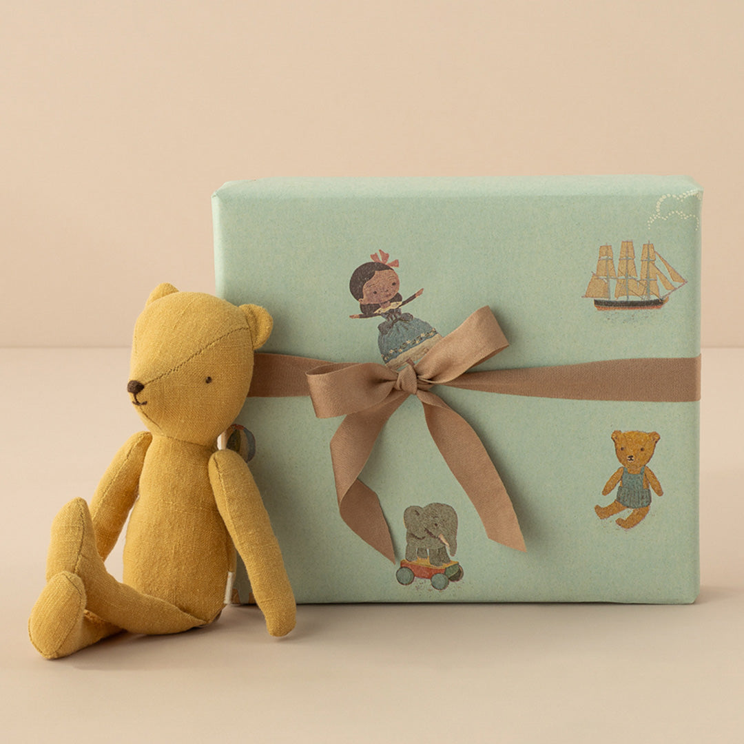 Een ingepakt cadeau met Maileg inpakpapier en een bruine strik, naast een geel stoffen beertje.