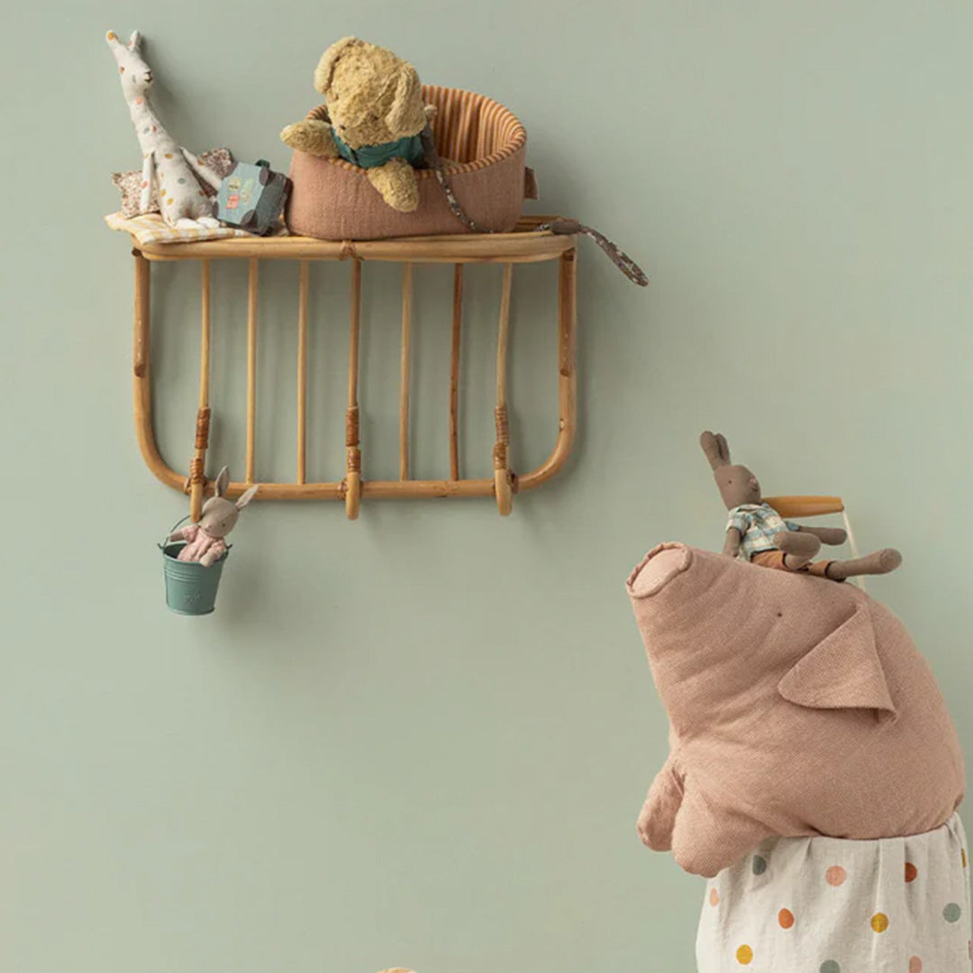 De Maileg knuffel puppy zit in een mandje op een houten wandplank, omringd door andere knuffels in een kinderkamer.