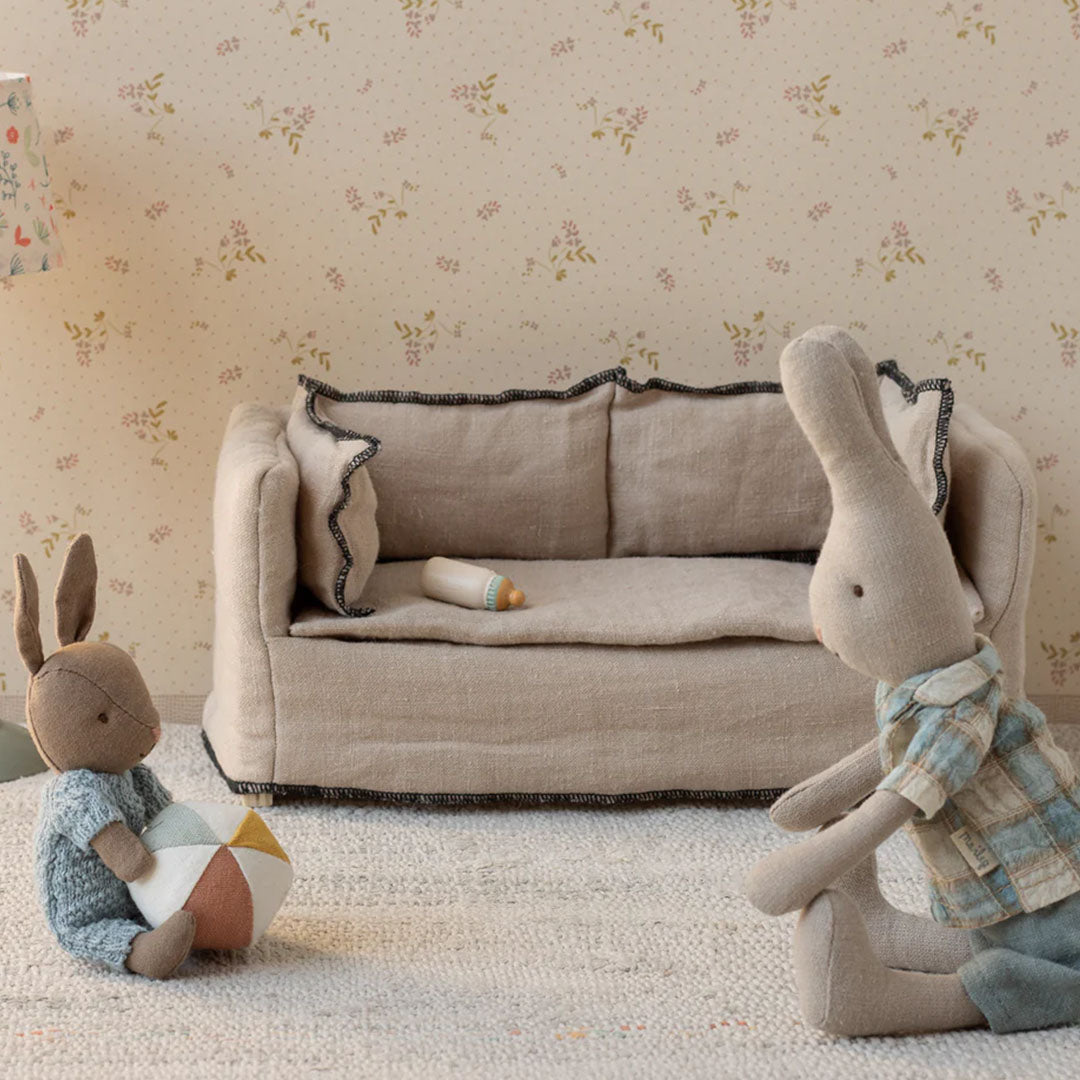 Twee Maileg pluchen konijnen spelen in een miniatuurkamer met een bankje en een flesje.