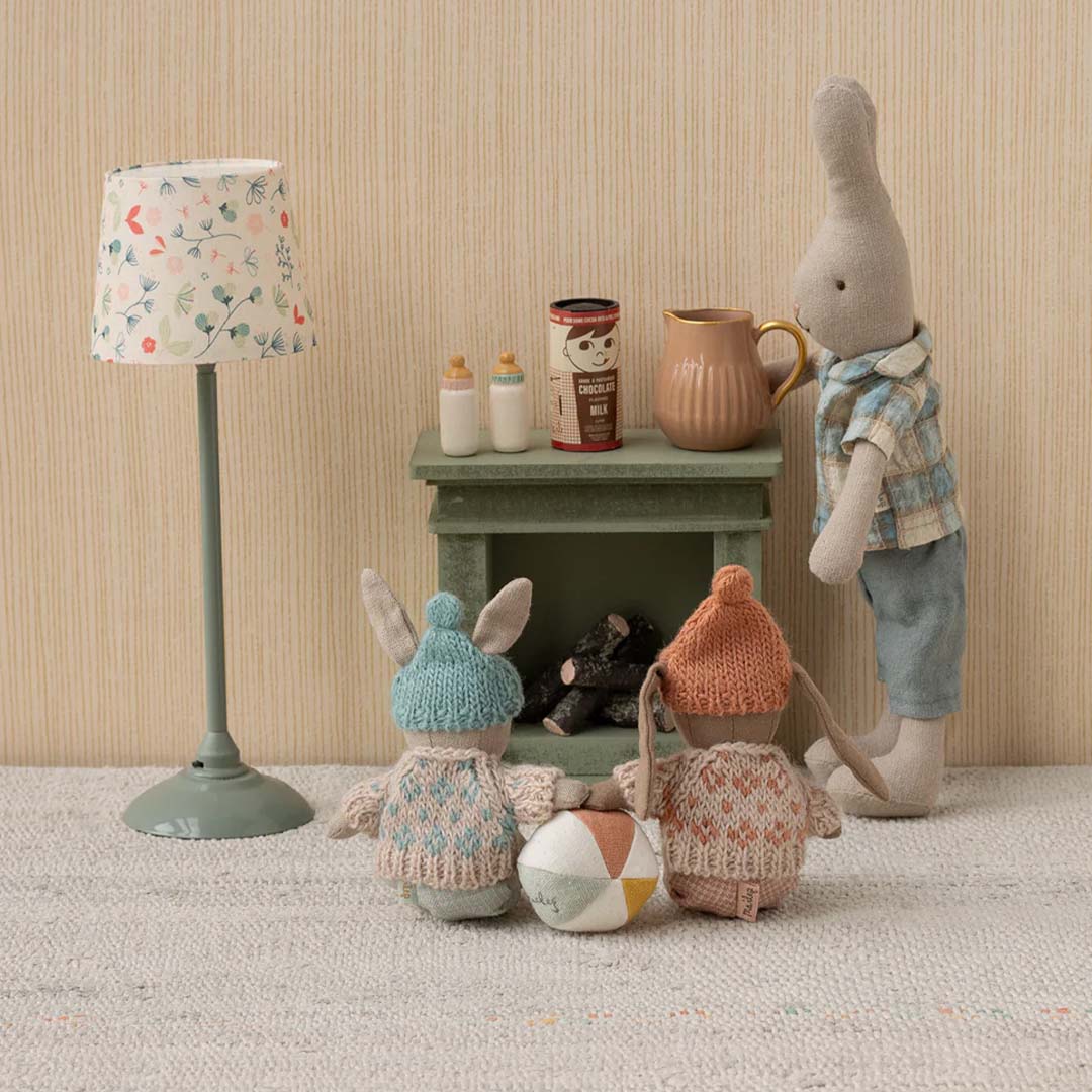 Drie Maileg knuffelkonijnen in een miniatuurkamer, waarvan twee met gebreide kleding op de rug gezien.