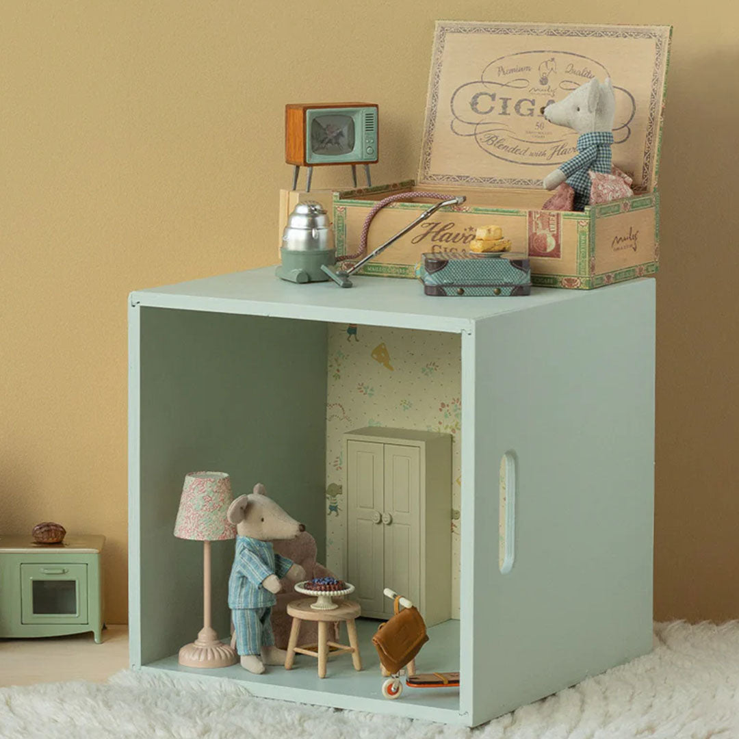Een Maileg knuffelmuis staat in een poppenhuis setting met meubels, terwijl een andere muis in de sigarendoos erboven zit.