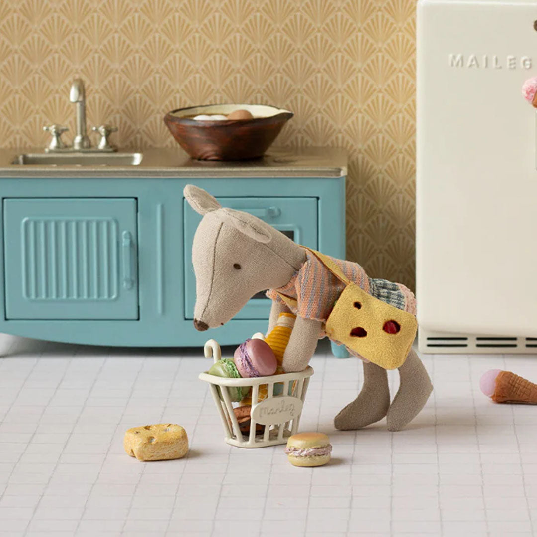 Maileg muis pop met gestreepte top en kaasvormige tas, die een boodschappenmandje met miniatuurgebak vasthoudt in een speelgoedkeuken.