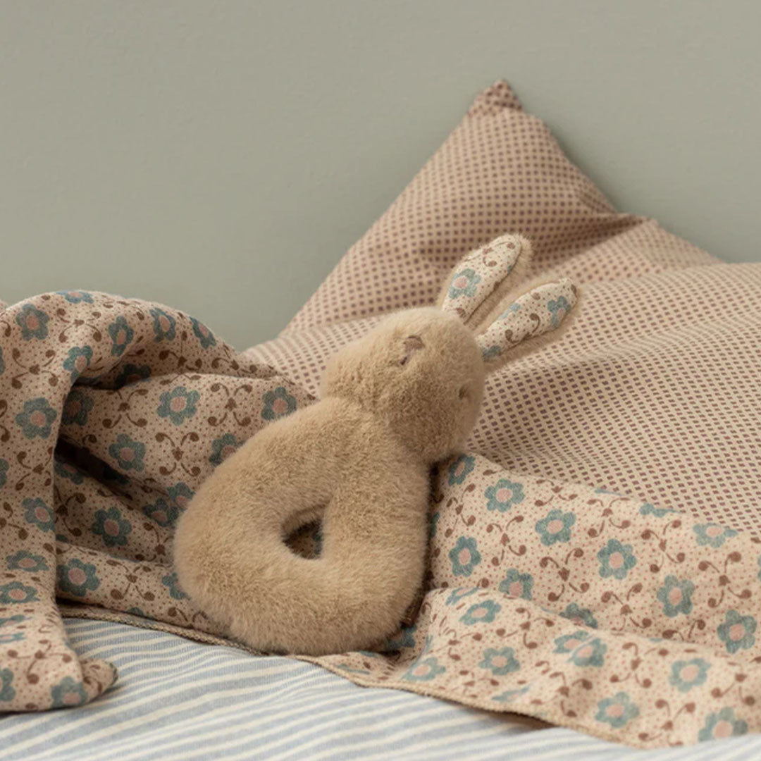 Een Maileg beige pluche konijnenrammelaar ligt op een gebloemd babydekentje.
