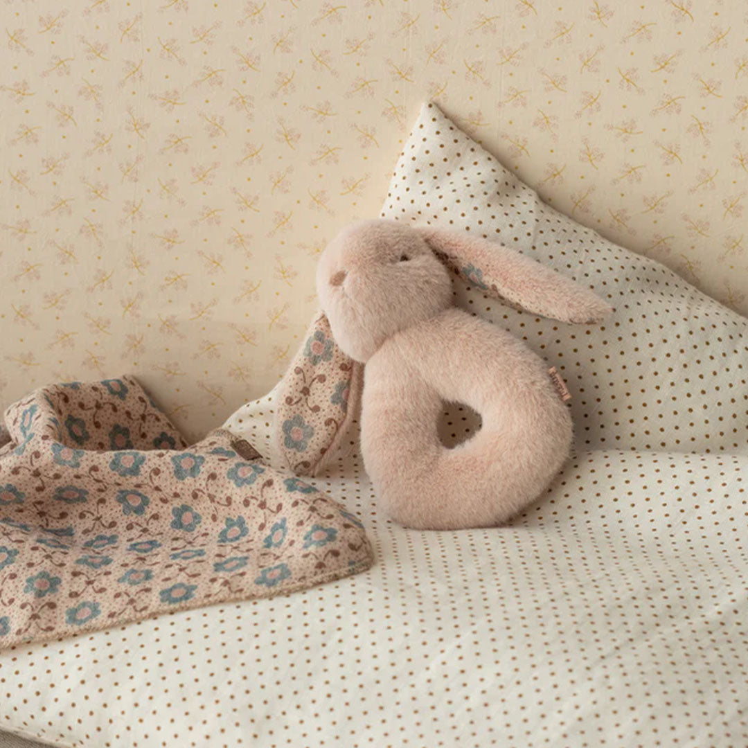 Een roze pluche konijnenrammelaar en een gebloemd babydekentje liggen op een bed met stippen.