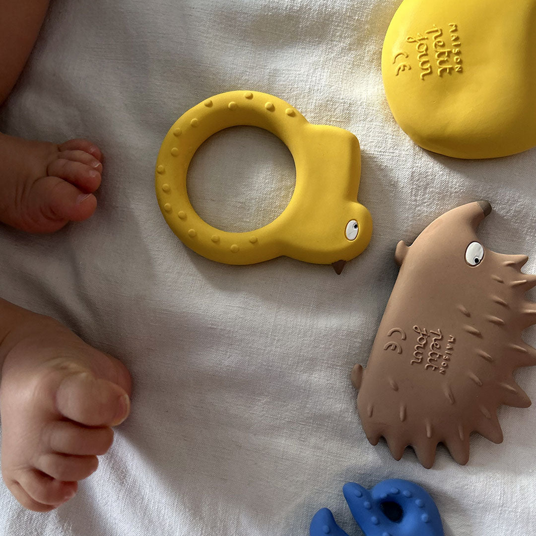 Babyvoetjes naast Maison Petit Jour bijtspeelgoed, waaronder de beige egel en een geel ringvormig speeltje, op een witte ondergrond.