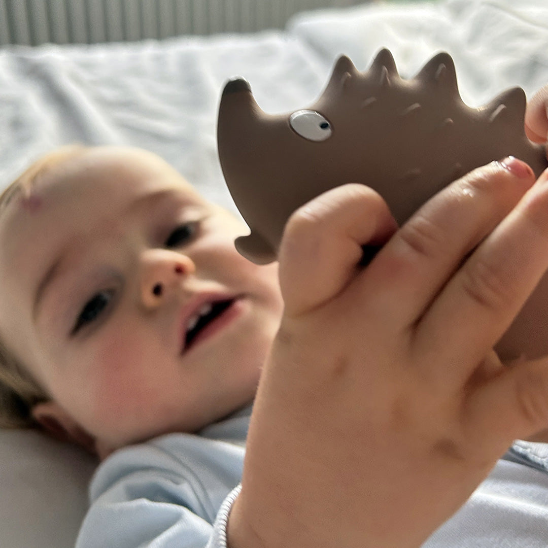 Een baby ligt op de rug en houdt het Maison Petit Jour beige egel bijtspeelgoed vast.