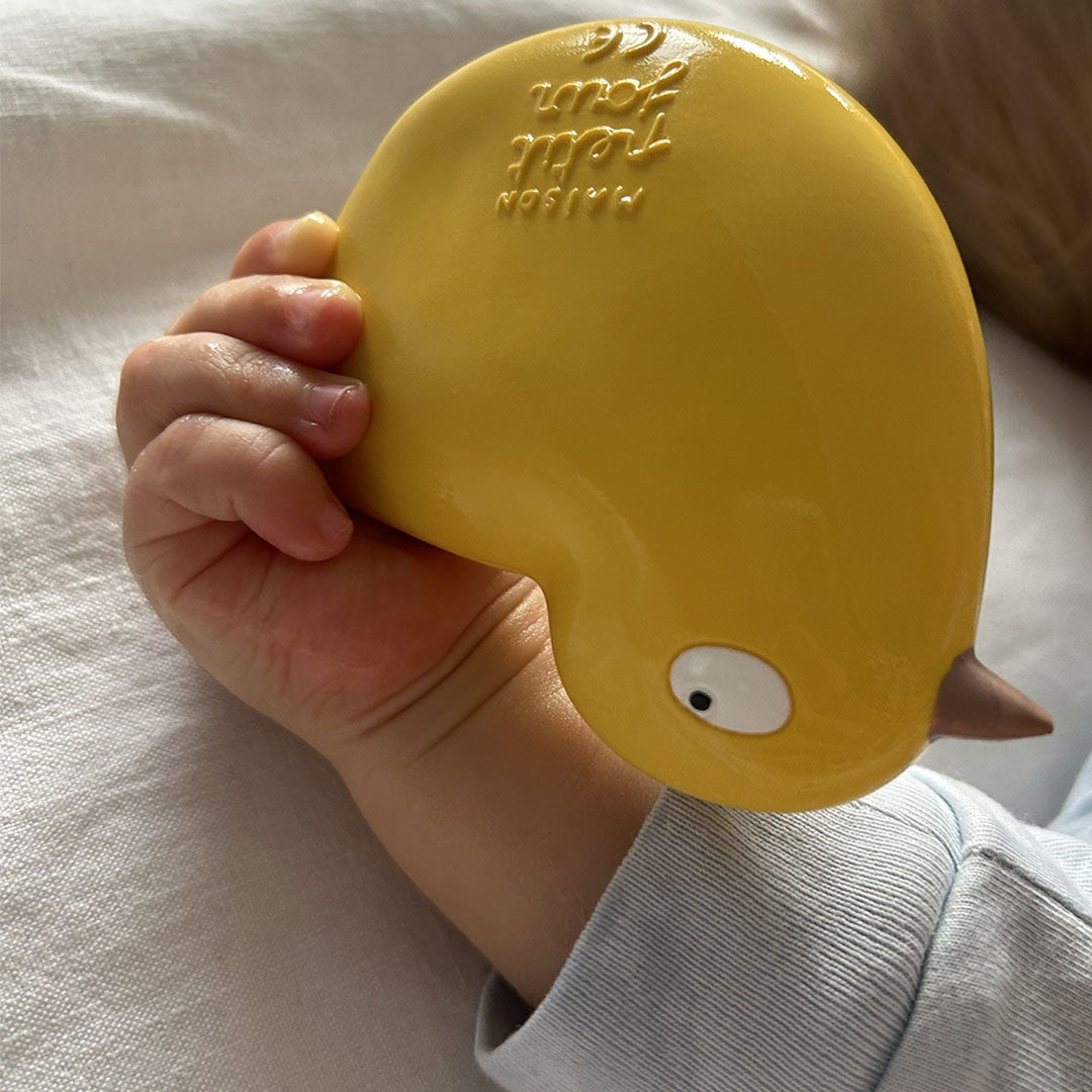Een babyhandje houdt het gele Maison petit jour rubberen bad- en bijtspeelgoed kuiken vast.