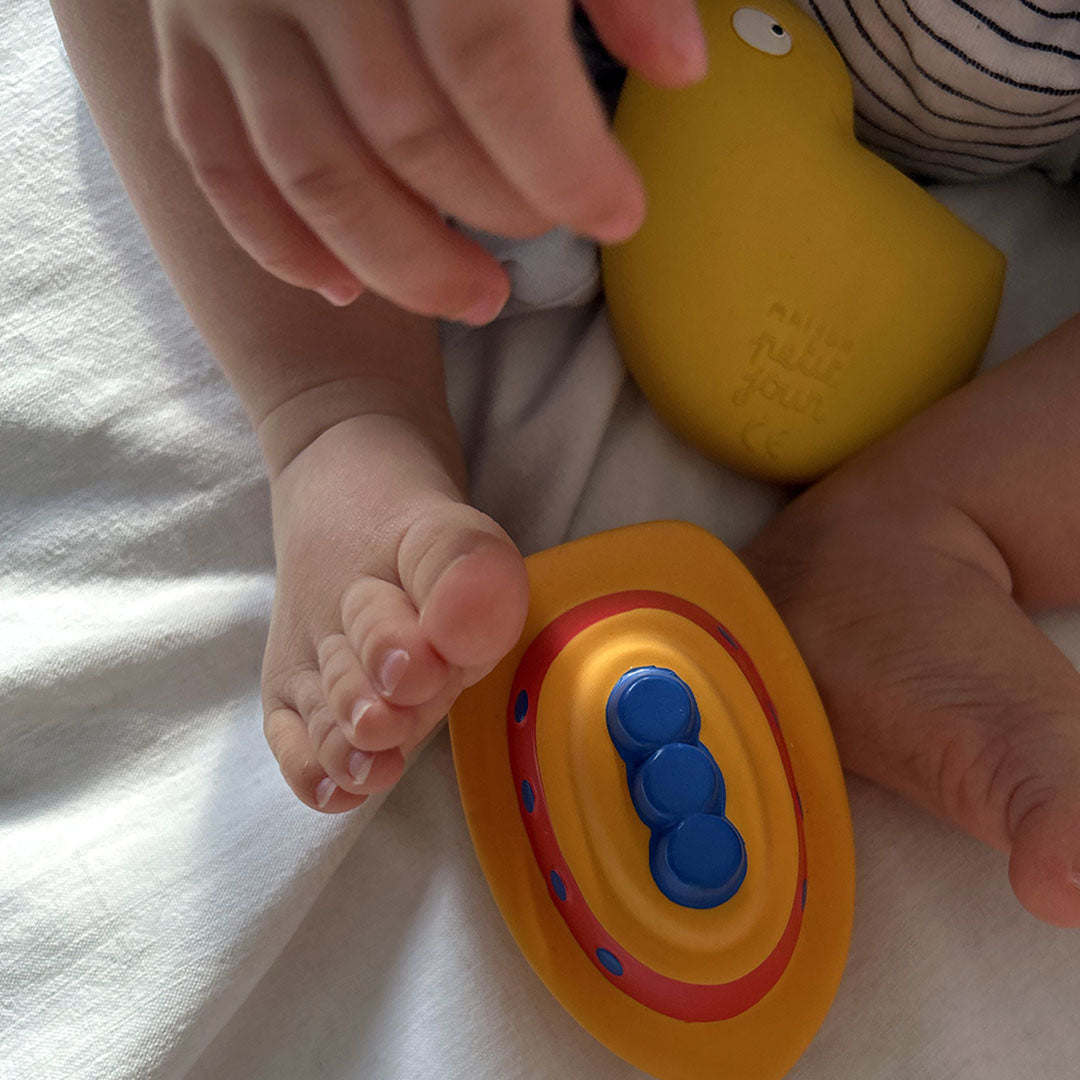 Babyvoetjes liggen naast het gele Maison petit jour bad- en bijtspeelgoed kuiken en een oranje bootje.