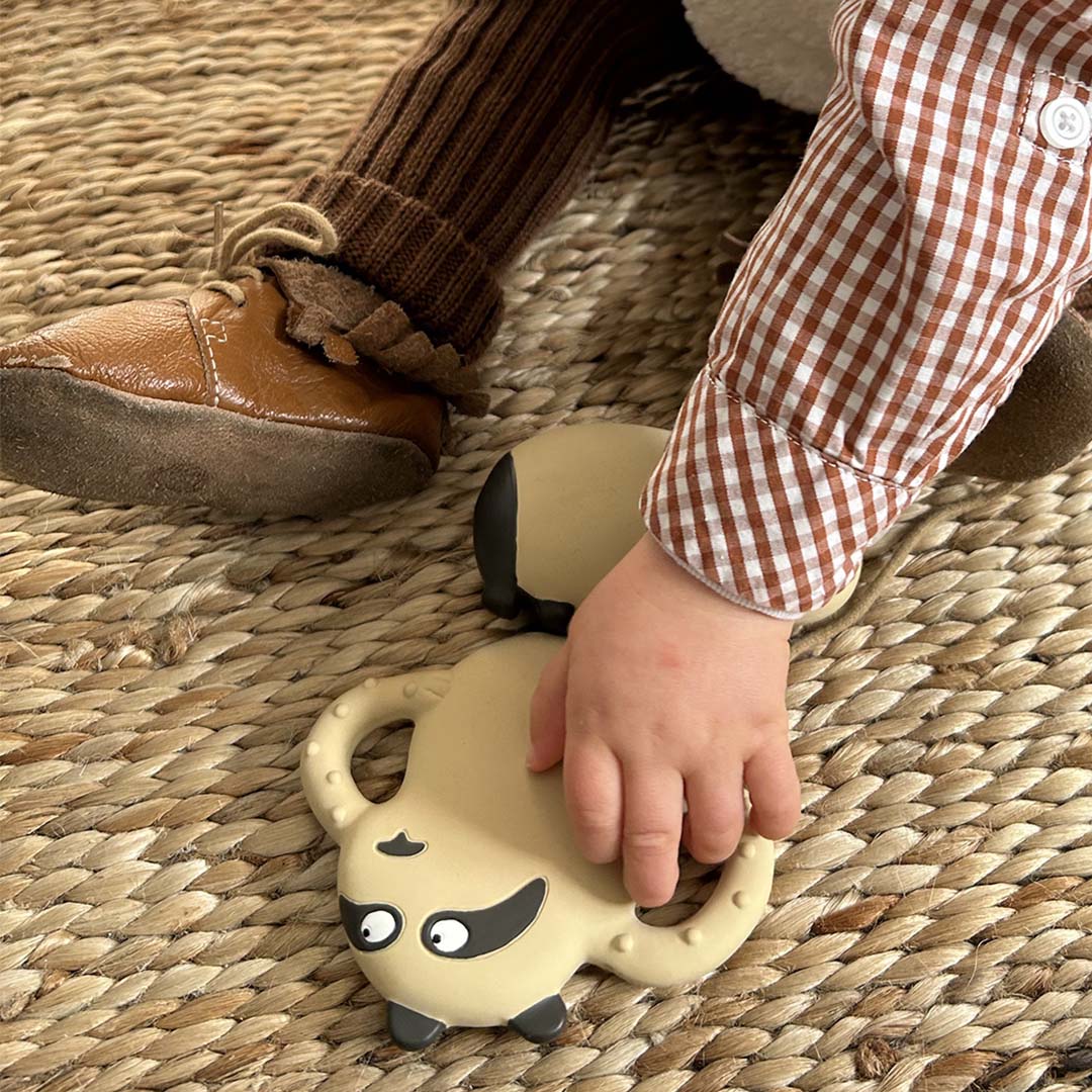 Een babyhandje reikt naar de Maison Petit Jour bijtring in de vorm van een beige panda op een mat.