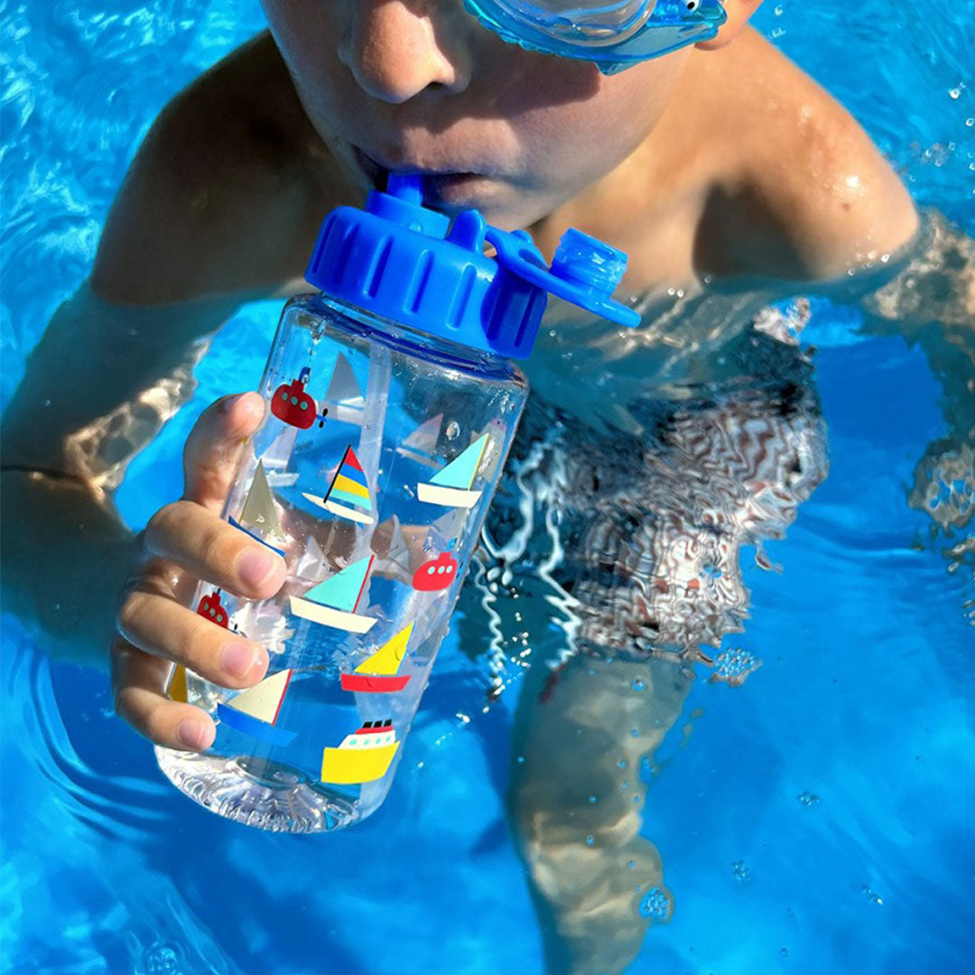 Een kind met blauwe duikbril drinkt uit de drinkfles met botenpatroon in een zwembad.