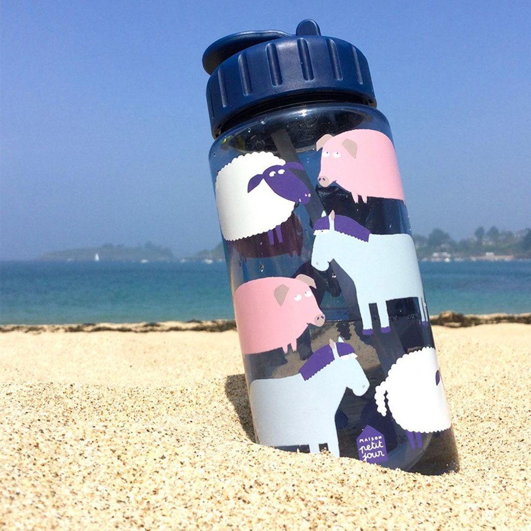 Maison Petit Jour kinder drinkfles met boerderijdieren patroon, staand in het zand op het strand.