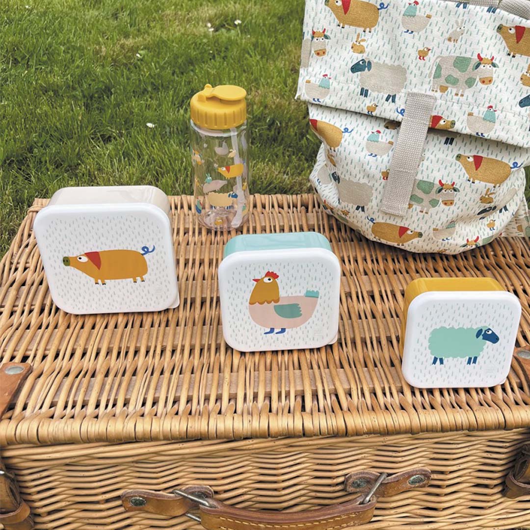 Maison petit jour drinkfles en drie lunchboxen met boerderijdieren op een picknickmand in het gras.