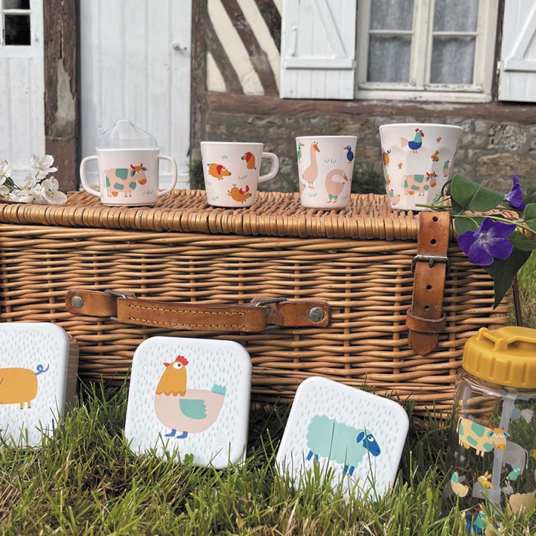Maison petit jour drinkfles en vier bekers met boerderijdieren op een picknickmand voor een huis.