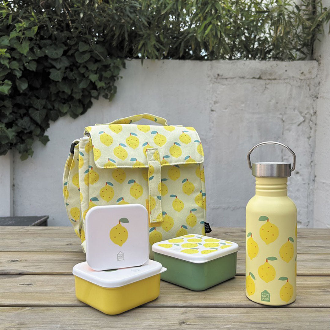 Een maison petit jour lunchset met gele lunchtas, waterfles en twee snackdoosjes met citroenprint op een houten tafel.