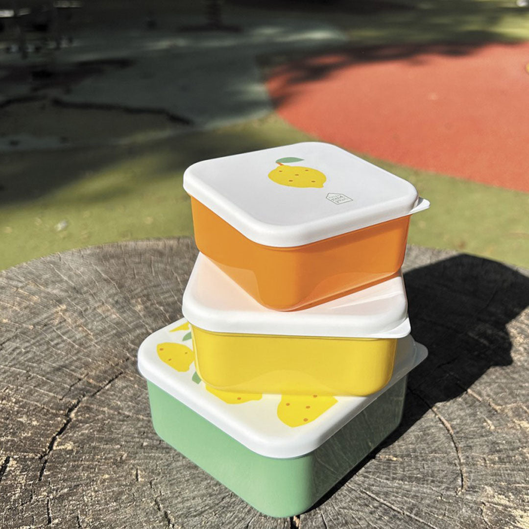 Drie gestapelde maison petit jour snackdoosjes in groen, geel en oranje met citroenprint op een boomstronk.