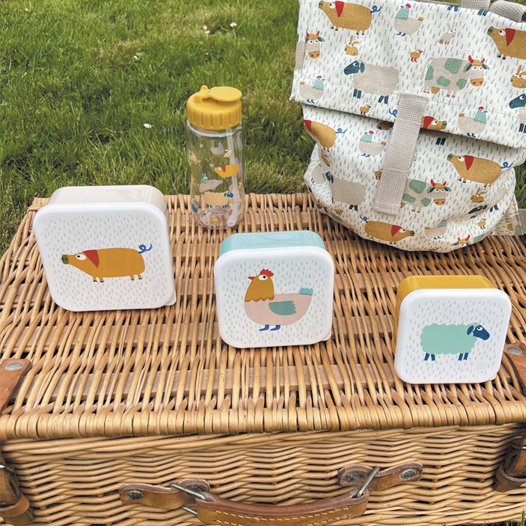 Maison Petit Jour snackdoosjes met boerderijdieren, een waterfles en rugzak op een picknickmand in het gras.