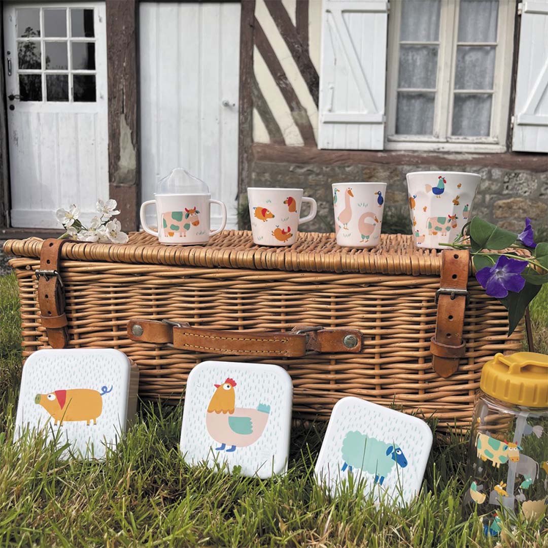 Maison Petit Jour snackdoosjes, waterfles en bekers met boerderijdieren op een picknickmand in het gras voor een huis.