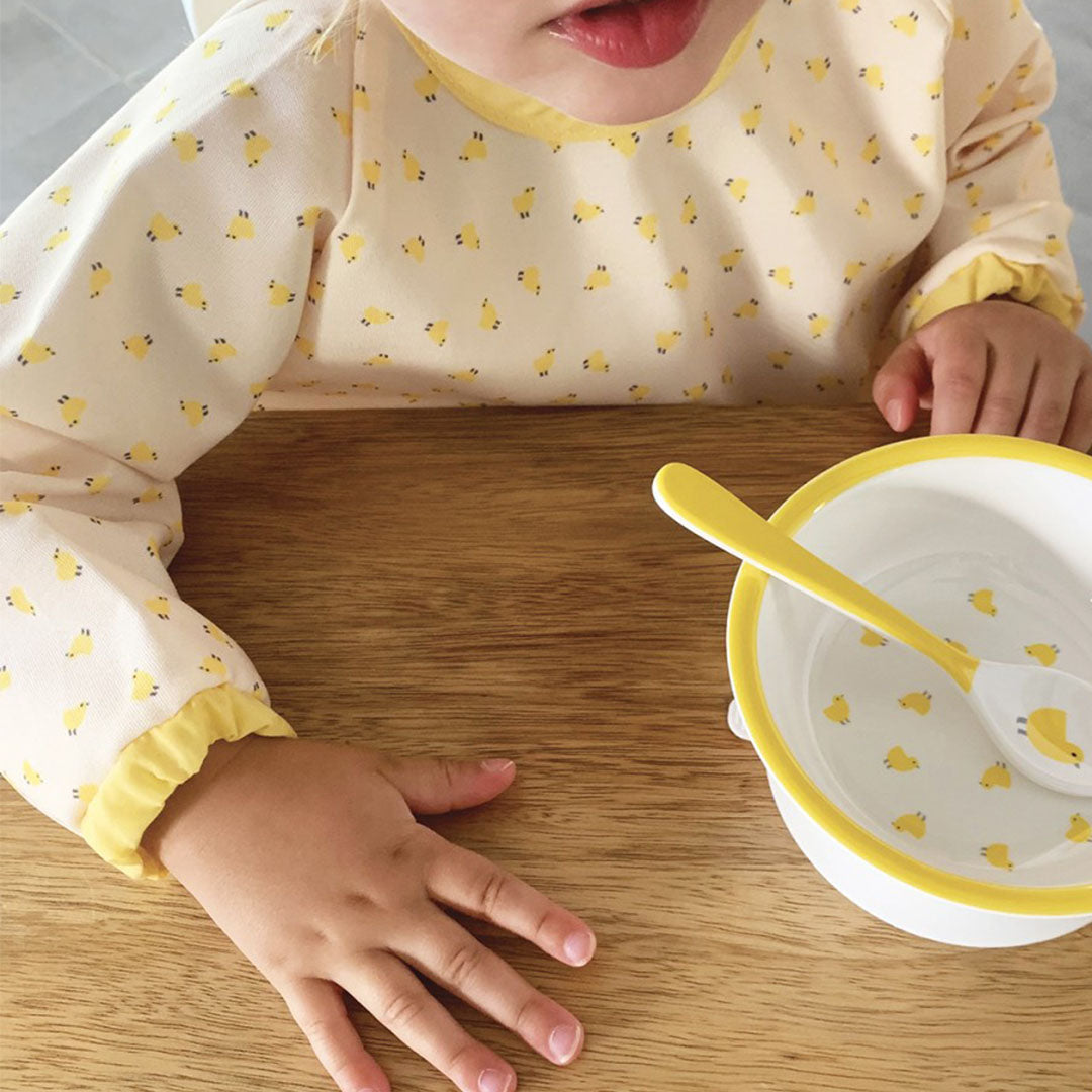 Baby draagt een mouwslab met kuikenpatroon tijdens het eten aan een houten tafel.