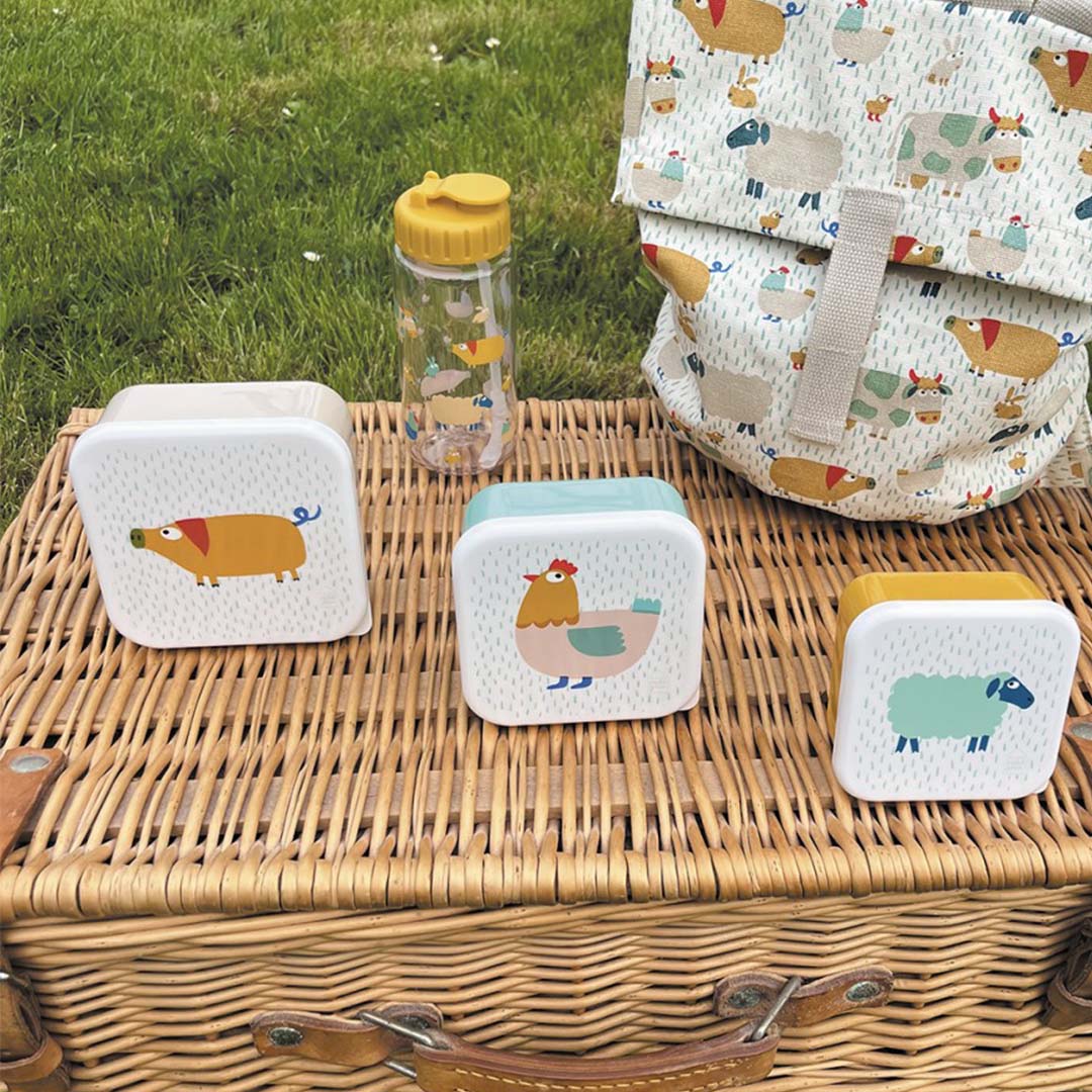 Maison Petit Jour schoolrugzak, drie lunchboxen en een drinkfles met boerderijdieren op een picknickmand in het gras.