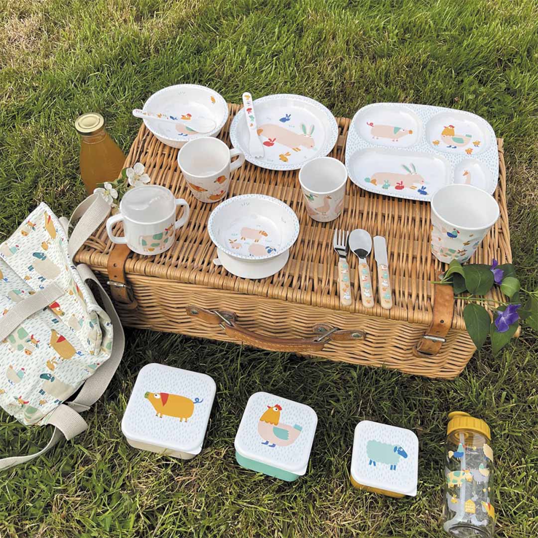 Maison Petit Jour schoolrugzak, kinderservies, lunchboxen en drinkfles met boerderijdieren op een picknickmand in het gras.