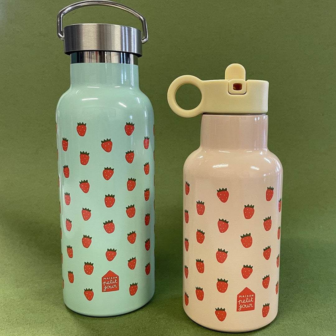 Twee Maison Petit Jour kinder drinkflessen met aardbeienprint, een roze en een lichtblauwe, staand op een groene ondergrond.