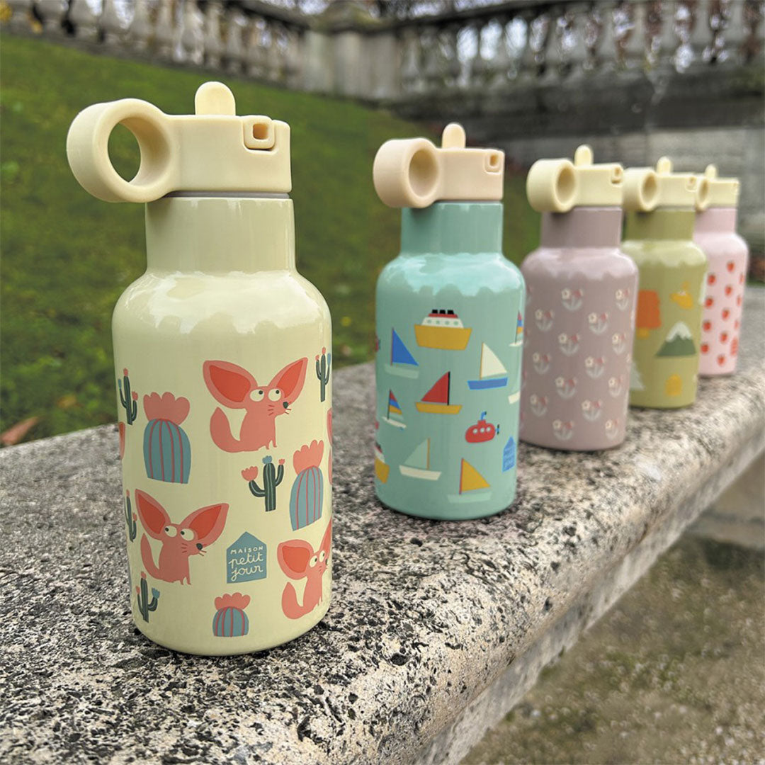 Rij van vijf Maison Petit Jour kinder drinkflessen met diverse kleurrijke patronen op een stenen muur.
