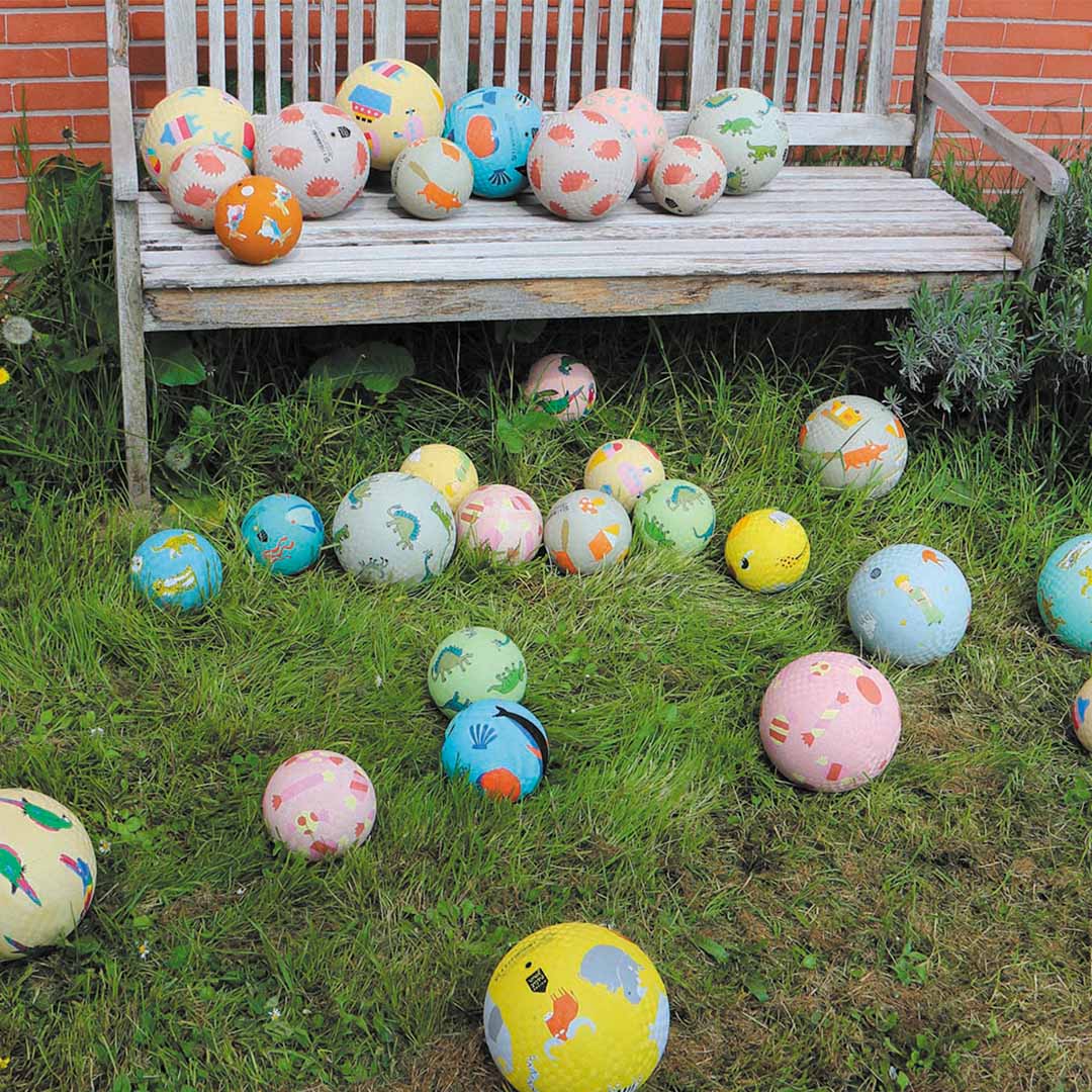 Een collectie van diverse gekleurde speelballen met dierenpatronen ligt op gras en een houten bank.