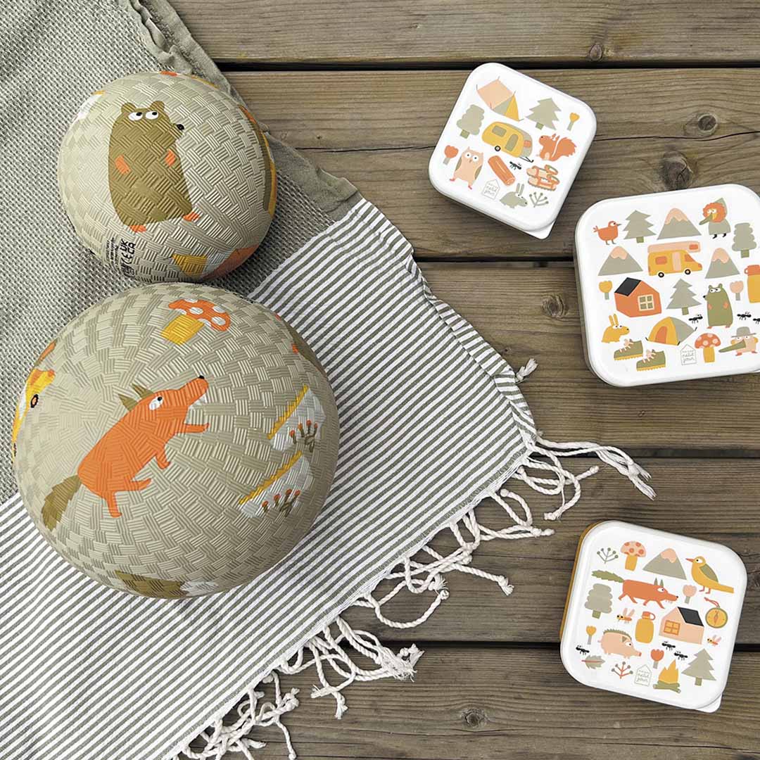 Twee Maison Petit Jour speelballen en drie bijpassende lunchboxen met avontuurlijke dieren en voertuigen op een houten tafel.
