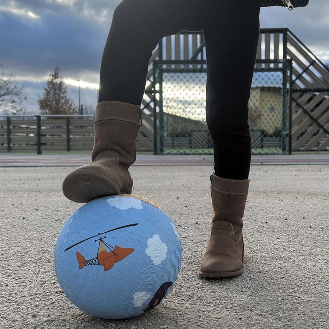 Een kind met bruine laarzen staat met één voet op een blauwe speelbal met een oranje helikopter op een grindpad.