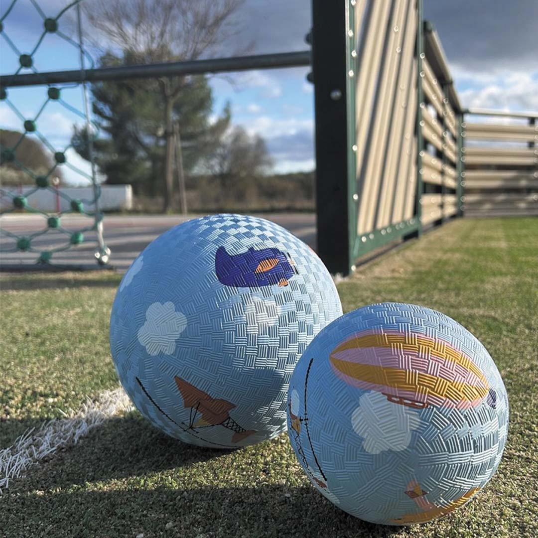 Twee blauwe speelballen met luchtvoertuigen en wolken liggen op groen gras naast een sportveld.