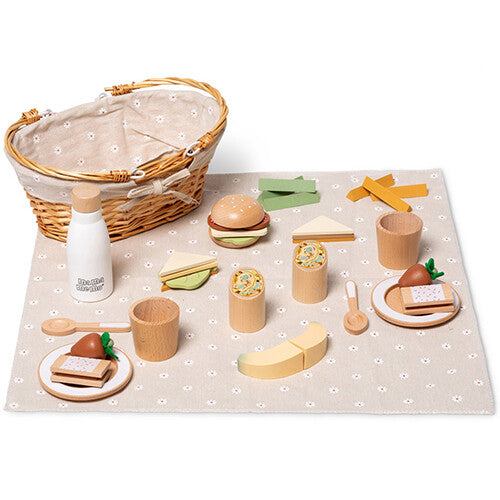 Een mama memo houten picknick speelset met mand, fles, bestek, borden en diverse speelgoedvoedsel zoals een burger, wraps en fruit, uitgestald op een gebloemd kleed.