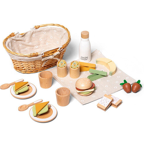 Een mama memo houten picknick speelset met mand, fles, bestek, borden en diverse speelgoedvoedsel zoals een burger, wraps, bananen en aardbeien, uitgestald op een gebloemd kleed.