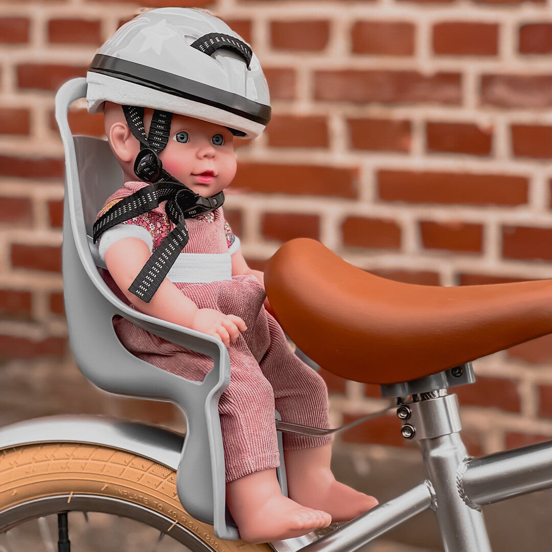 Een pop met helm zit in een mama memo poppenfietszitje op een fiets voor een bakstenen muur.