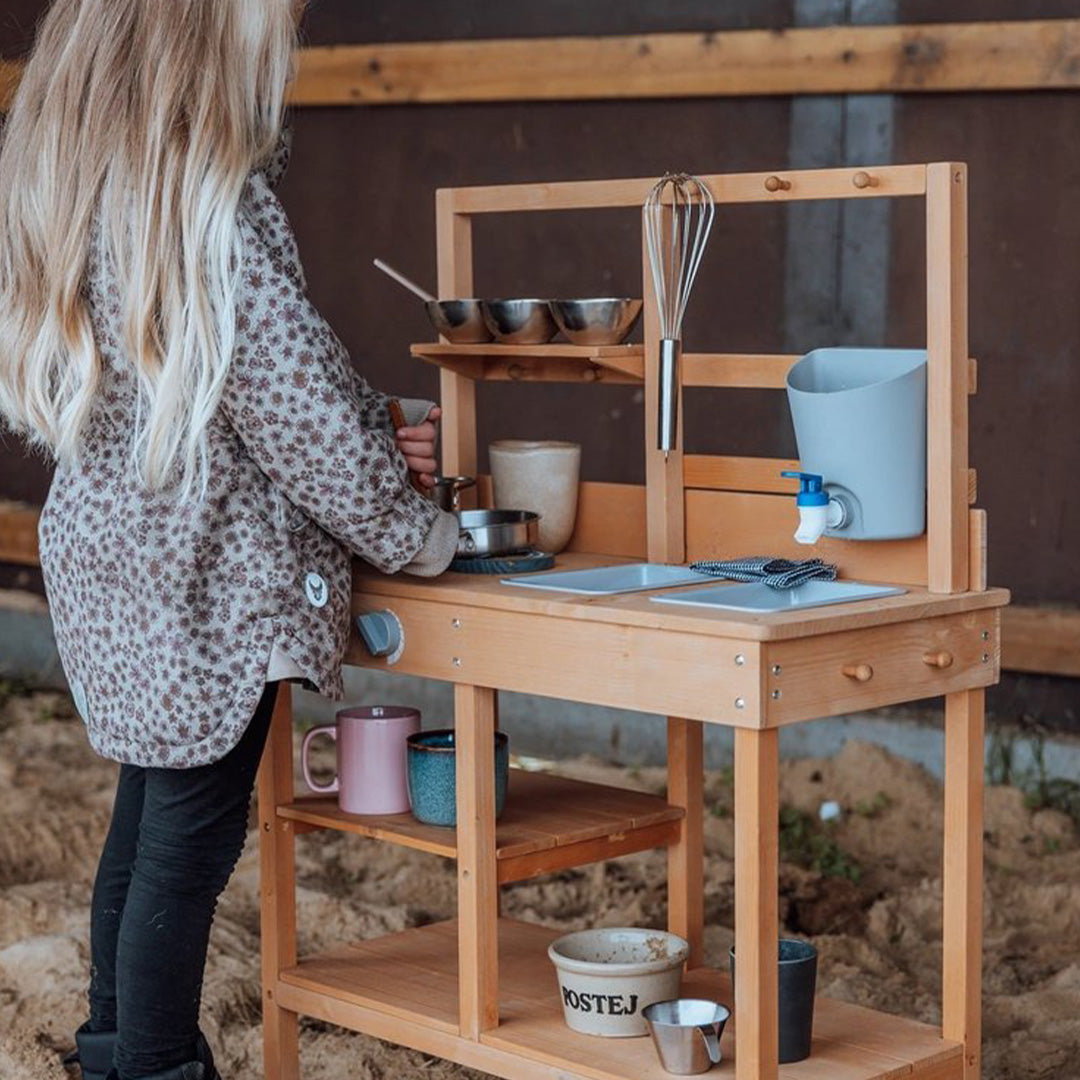 Een kind speelt met de mama memo houten kinder buitenkeuken in een zanderige omgeving.